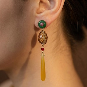 Peut inclure: Boucle d'oreille pendante avec un clou vert et un centre rose. La boucle d'oreille comprend un ovale marron tacheté, une perle rouge et un pendentif jaune en forme de goutte. La boucle d'oreille est dorée et pend près de l'oreille d'une personne.