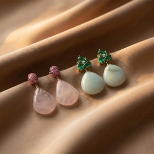 May include: Two pairs of gemstone earrings displayed on a tan fabric. One pair features pink teardrop-shaped stones with pink gem tops. The other pair has green gem tops and light green teardrop-shaped stones. Jewellery.