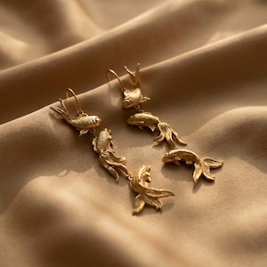 May include: Gold-coloured fish earrings. Each earring has a hook and a series of detailed fish charms, creating a dangling effect. The fish have textured scales and flowing fins, adding to their realistic appearance.