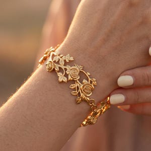 May include: A gold bracelet with a floral design, featuring roses and leaves. The bracelet is made of a shiny metal and is connected by a chain. The bracelet is worn on a wrist.