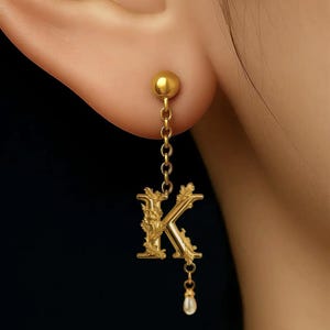 May include: Gold-colored earring featuring the letter "K" in an ornate, floral design. The earring hangs from a small chain and is accented with a dangling pearl. The earring is shown on a person's ear.