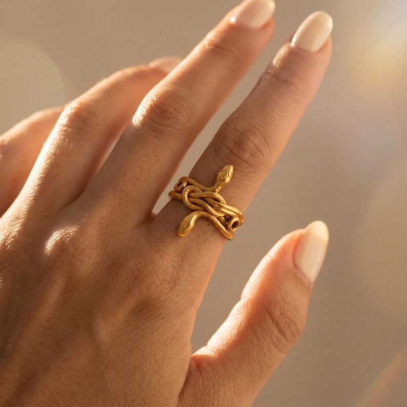 May include: A gold-colored ring featuring a snake design. The ring is wrapped around the finger, with the snake's head and tail visible. The ring is made of a shiny, metallic material.