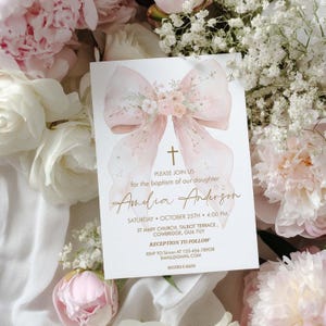 Puede incluir: Una tarjeta de invitación de bautizo con un diseño de lazo y cruz en acuarela. La tarjeta presenta el nombre "Amelia Anderson" en una escritura elegante, junto con los detalles del evento. Se utilizan tonos rosas y dorados suaves, rodeados de flores.