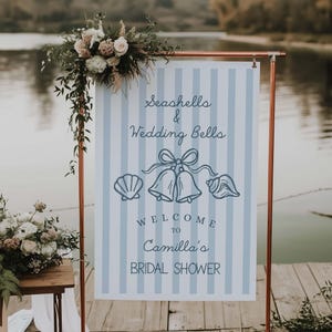 May include: A sign with blue and white stripes, copper frame, and floral arrangement. The sign reads "Seashells & Wedding Bells" and "Welcome to Camilla's Bridal Shower" with seashell and bell illustrations.
