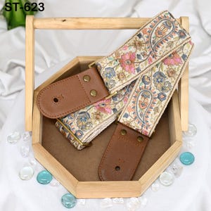 May include: An embroidered guitar strap with a paisley floral design in pink, blue, and beige. The strap has brown leather ends and gold-colored hardware. The text "ST-623" is in the upper left corner.