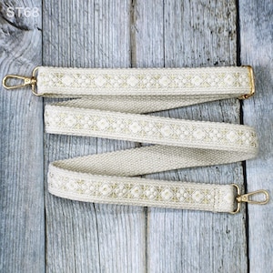 May include: Cream-colored bag strap with gold-colored hardware. The strap features a woven texture with a repeating floral and geometric pattern in gold. The strap has gold-colored metal clasps on each end for easy attachment to a bag.