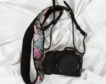 以下が含まれることがあります： 黒いニコンカメラと花の刺繍入りカメラストラップ。ストラップは、黒い背景にピンク、緑、白の花が特徴です。「ST-703」の文字が左上にあります。