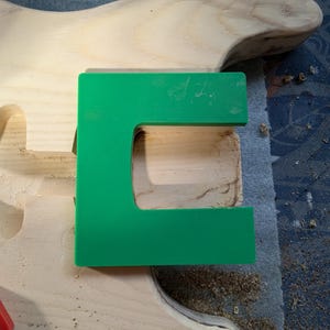 Op de afbeelding: Een close-up van een houten gitaarlichaam met een groene plastic sjabloon erop. De sjabloon is C-vormig en wordt gebruikt om het uitsnijden van het gitaarlichaam te begeleiden.