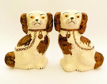 May include: Pair of ceramic dog figurines, each with white fur, brown markings, and a gold chain collar with a padlock. The dogs are sitting with their heads tilted down and their tails curled.