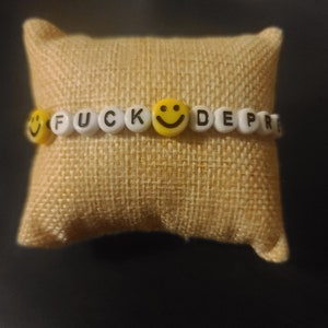 May include: A white beaded bracelet with the words "F**K DEPRESSION" and a yellow smiley face charm.