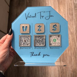 以下が含まれることがあります： 3つの支払いアイコンとQRコードが付いた、水色のバーチャルチップジャー。テキスト「Virtual Tip Jar」と「Thank you」が表示されています。ジャーは透明なアクリルベースです。