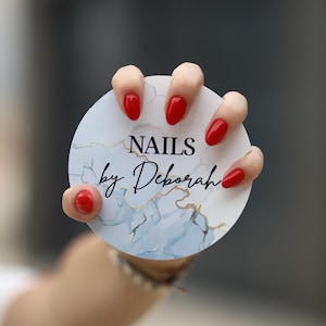 Puede incluir: Una tarjeta redonda blanca con un diseño de mármol azul y dorado. La tarjeta tiene el texto "NAILS by Deborah" en escritura cursiva negra. Una mano con esmalte de uñas rojo sostiene la tarjeta.