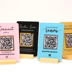 Peut inclure: Quatre panneaux acryliques avec des codes QR et du texte. Les panneaux sont rose, noir, bleu et or. Le texte sur les panneaux comprend "Connect with us", "Emilia Sion Editorial Services", "SCAN TO PAY BY Venimo", et "Scan to Book an Appointment".