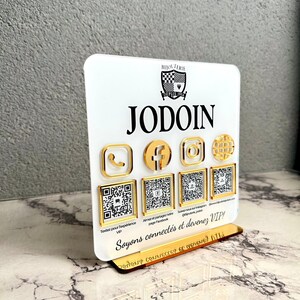 May include: A white square acrylic sign with a gold base featuring the black text BIJOUTERIE DEPUIS 1954 JODOIN. It displays four gold social media icons above four square QR codes, with the text Soyons connectés et devenez VIP! at the bottom.