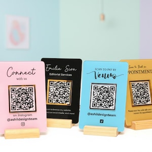 以下が含まれることがあります： ピンク、黒、青、金色の4色のQRコード名刺。それぞれにユニークなデザインとテキストが施されています。カードは小さな木製のスタンドに表示され、「Connect with us」や「Scan to pay by Venmo」などのテキストが表示されています。