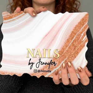 Könnte beinhalten: Ein rechteckiges Schild mit gewellten Kanten, das ein weißes und blassrosa Marmormuster mit diagonalen Bändern aus Kupferglitzer aufweist. Der Text NAILS by Jennifer ist mittig in Gold und Schwarz gedruckt, darunter das Instagram-Symbol und @Jenails.