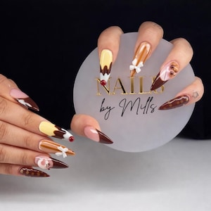 Könnte beinhalten: Lange, spitze Acrylnägel mit Dessert-Motiven. Designs beinhalten Schokoladen-, Karamell- und Vanilleeis, mit weißen Schleifen und roten Akzenten. Die Nägel werden vor einem Kreis aus mattiertem Glas mit dem Text "NAILS by Mills" in Gold gehalten.