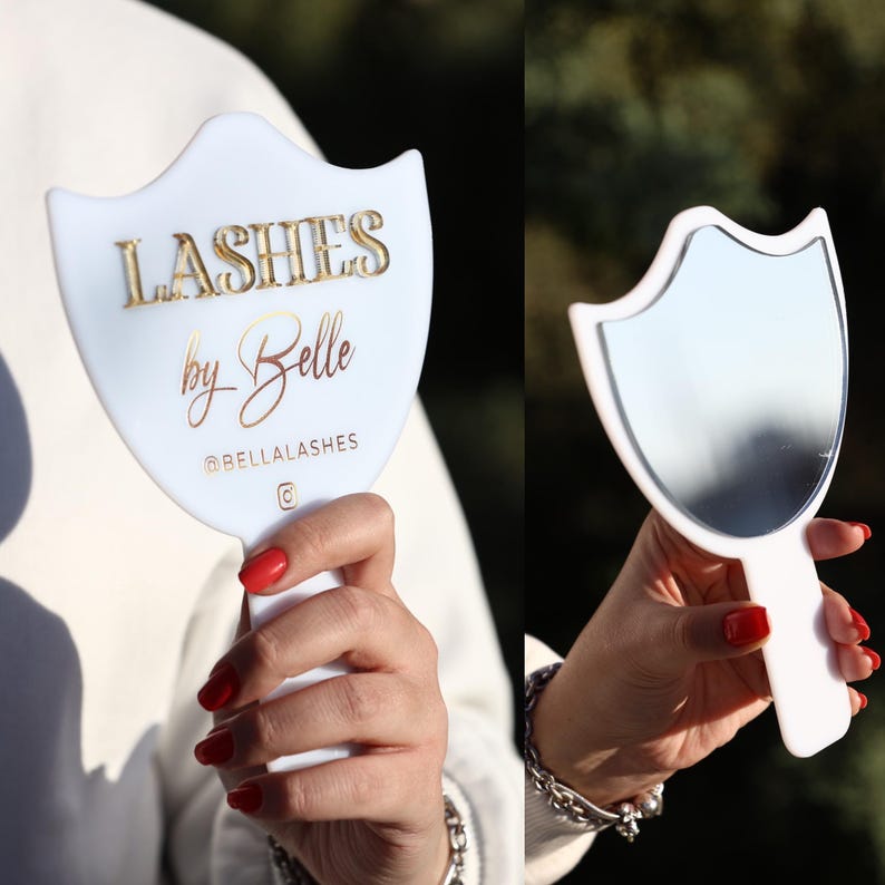 May include: White hand mirror with a shield-shaped design. The mirror has a gold inscription on it