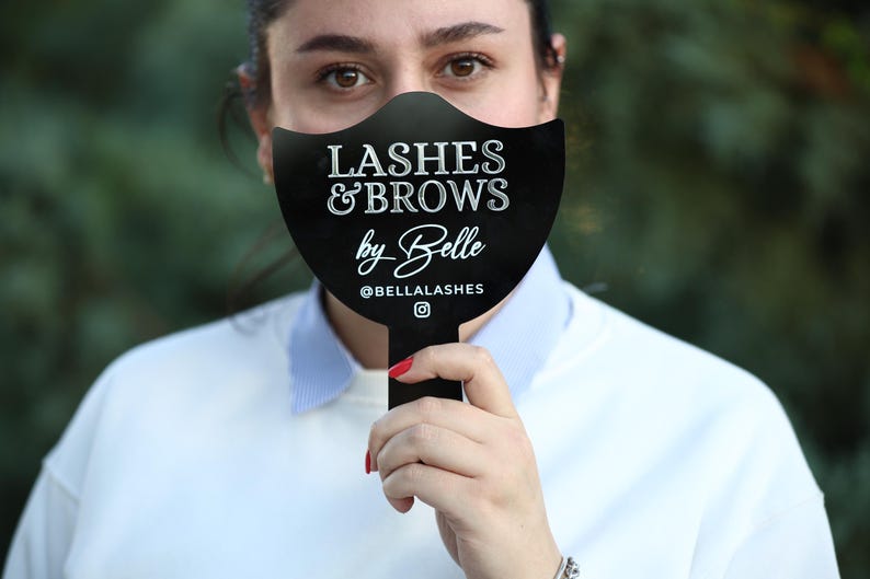May include: A black sign with white text that reads "Lashes & Brows by Belle @BELLALASHES" with a small Instagram icon. The sign is held in front of a person's face.
