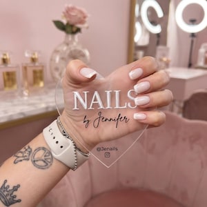 Könnte beinhalten: Ein transparentes, herzförmiges Acrylschild mit dem weißen Schriftzug NAILS und dem schwarzen Schreibschrifttext by Jennifer, ergänzt durch den Social-Media-Namen @Jenails und ein Instagram-Symbol am unteren Rand zur Markenpräsentation.