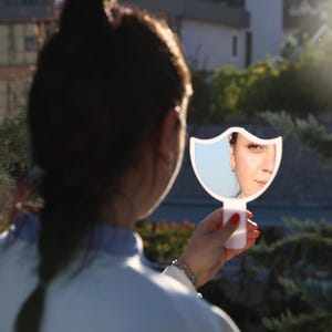 May include: A woman looks into a white hand mirror with a bird-shaped frame. The mirror reflects her face and the background behind her.