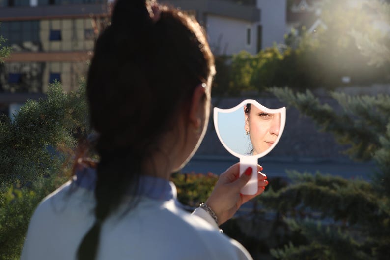 May include: A woman looks into a white hand mirror with a bird-shaped frame. The mirror reflects her face and the background behind her.