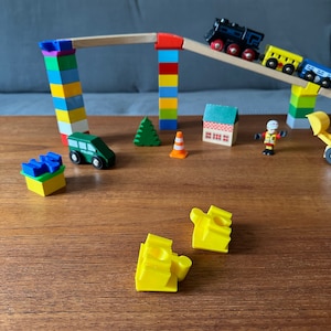 Könnte beinhalten: Ein hölzernes Spielzeug-Zugset mit einer schwarzen Lokomotive und bunten Waggons fährt eine Holzrampe hinunter, die von bunten Blöcken getragen wird. Weitere Holzspielzeuge sind ein grünes Polizeiauto, ein Haus, ein Baum und ein Bauarbeiter.