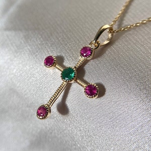Puede incluir: Un collar con un colgante de cruz de oro con cinco piedras preciosas. La piedra central es una esmeralda verde, y las cuatro piedras que la rodean son rubíes rosas.