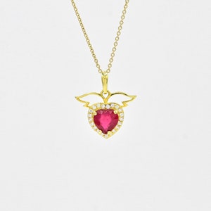 May include: A gold necklace with a heart-shaped pendant. The pendant has a bright red gemstone at its centre, encircled by small, clear stones. Gold-coloured wings are attached to the top of the heart, suspended from a delicate chain.