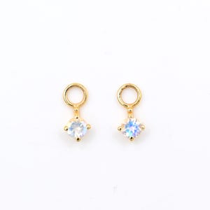 May include: Two gold-colored earrings with a small, round loop at the top and a square, iridescent gemstone at the bottom. The gemstones have a white and blue color. The earrings are set against a white background.