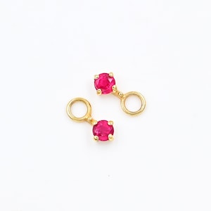 May include: Two gold-coloured jewellery pieces, each featuring a vibrant red gemstone and a circular loop. The gemstones are round and set in a four-prong setting. The pieces are displayed against a white background.