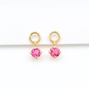 May include: A pair of gold-colored earrings, each featuring a bright pink gemstone. The gemstones are round and set in a four-prong setting. The earrings have a small loop at the top for attachment.