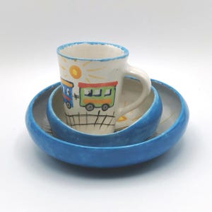 Könnte beinhalten: Ein handgefertigtes Keramik-Set mit einer Tasse und zwei Schalen. Die Tasse zeigt ein farbenfrohes Zugdesign mit Sonne und Bahngleisen. Die Schalen sind blau, die Tasse steht in der größeren Schale.