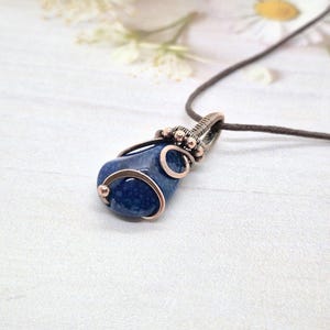Peut inclure: Pendentif artisanal avec une pierre bleu foncé, fil enroulé en cuivre. La pierre est de forme arrondie et irrégulière, avec une texture tachetée. Le pendentif est suspendu à un cordon marron. Le design est simple mais élégant.