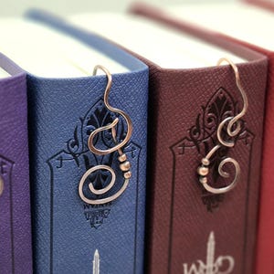 Könnte beinhalten: Nahaufnahme von vier Büchern mit dekorativen Kupferdraht-Lesezeichen. Die Bücher sind lila, blau, weinrot und rot. Jedes Lesezeichen hat ein einzigartiges Design, einschließlich Wirbeln und Buchstabenformen. Die Bücher haben geprägte Muster auf den Einbänden.