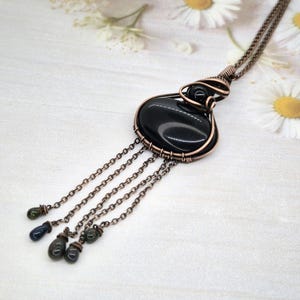 Peut inclure: Un collier pendentif artisanal avec une grosse pierre noire polie enveloppée dans du fil de cuivre. Le pendentif est suspendu à une chaîne délicate et orné de chaînes pendantes et de petites perles vertes et bleues foncées. Le collier est présenté sur une surface blanche.