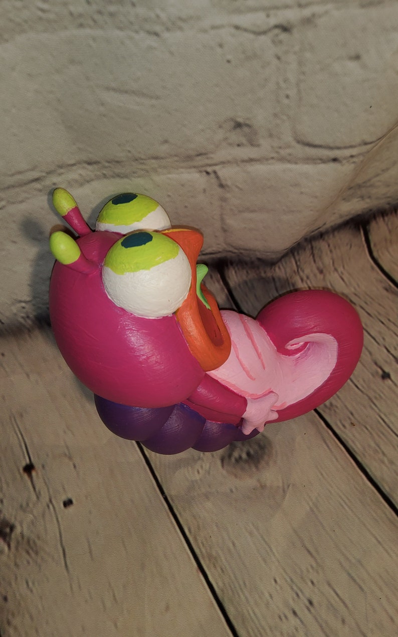 Op de afbeelding: Een roze en paarse cartoonrups speelgoed met grote groene ogen en een open mond. Het speelgoed is gemaakt van zacht, buigzaam materiaal en is in een speelse pose.