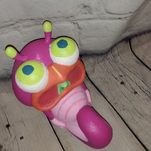 Op de afbeelding: Een roze en groene buitenaardse wezens met grote ogen en een wijd open mond. Het wezen is gemaakt van klei en is geschilderd met felle kleuren.