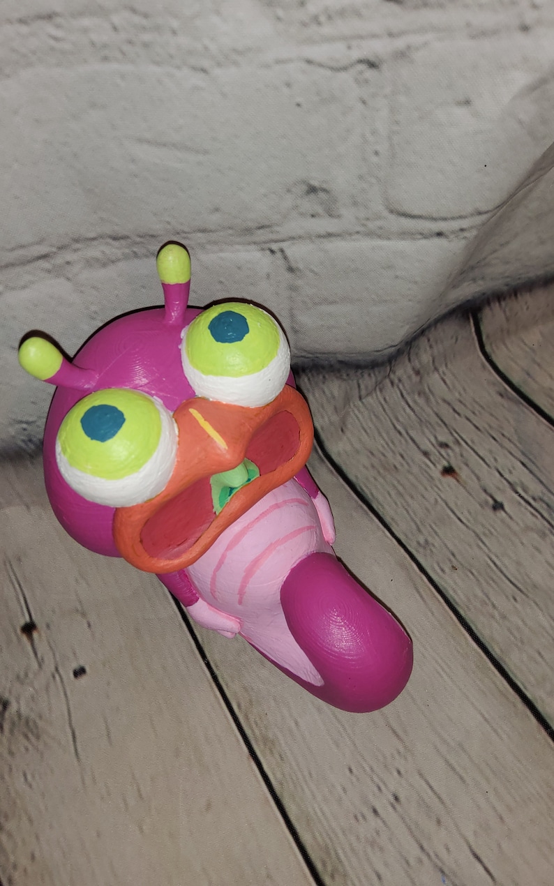 Op de afbeelding: Een roze en groene buitenaardse wezens met grote ogen en een wijd open mond. Het wezen is gemaakt van klei en is geschilderd met felle kleuren.