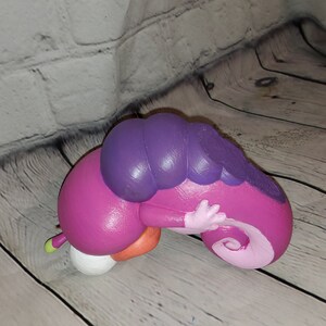 Op de afbeelding: Een roze en paarse opgerolde rupsfiguur met een witte buik. Het beeldje is gemaakt van geverfd keramiek en heeft een glanzende afwerking.