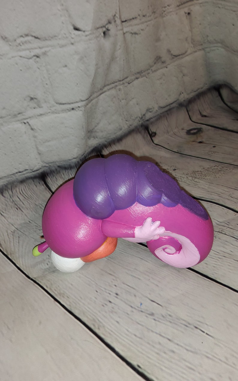 Op de afbeelding: Een roze en paarse opgerolde rupsfiguur met een witte buik. Het beeldje is gemaakt van geverfd keramiek en heeft een glanzende afwerking.