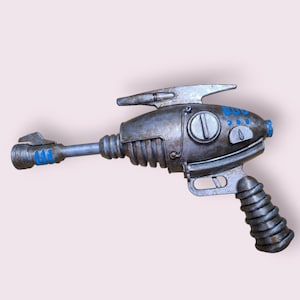 Puede incluir: Una pistola de rayos de juguete de color plateado con un diseño azul y blanco. La pistola tiene un mango y un cañón con una mira telescópica.