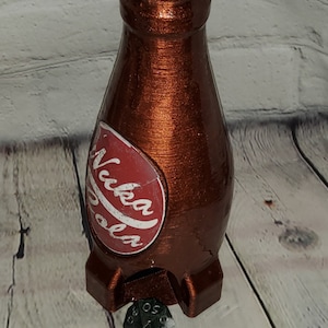 May include: A brown, 3D printed Nuka Cola bottle with a red and white label. The bottle is sitting on a wooden surface with a black and green dice in front of it.