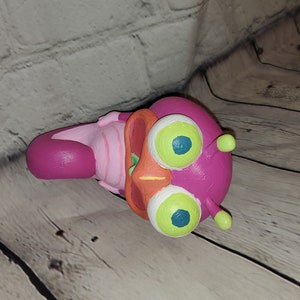 Op de afbeelding: Een handgeschilderd beeldje van roze en oranje kunststof, dat een cartoonfiguur met grote groene ogen voorstelt. Het figuur heeft een wijd open mond en een roze-wit gestreept lichaam.