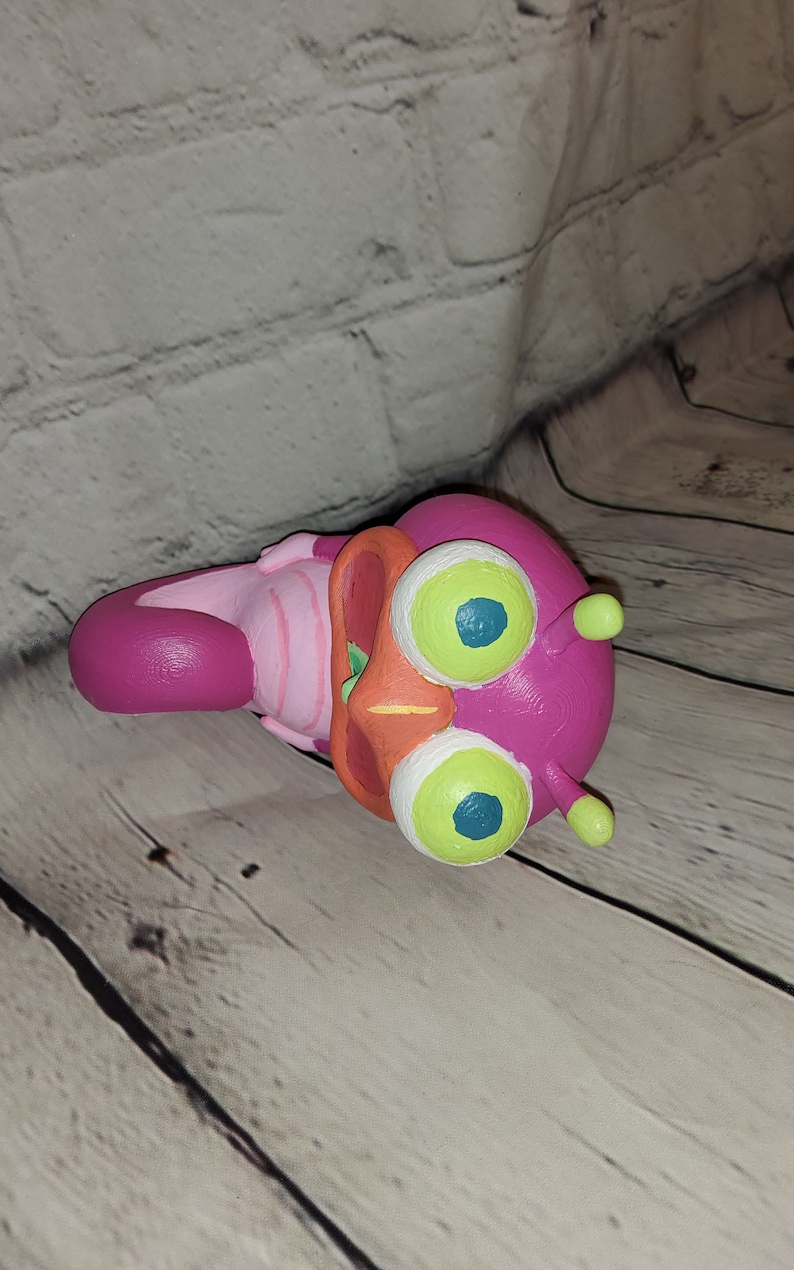 Op de afbeelding: Een handgeschilderd beeldje van roze en oranje kunststof, dat een cartoonfiguur met grote groene ogen voorstelt. Het figuur heeft een wijd open mond en een roze-wit gestreept lichaam.