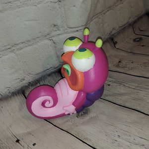 Op de afbeelding: Een roze en paarse cartoonfiguur met grote witte ogen en een groen en oranje mond. Het figuur heeft een gekrulde staart en is gemaakt van klei.