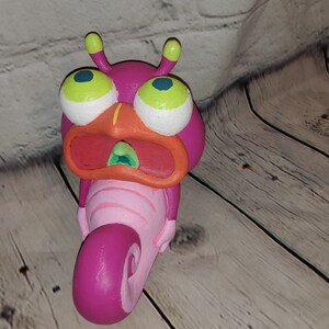 Op de afbeelding: Een handbeschilderd keramiek beeldje van een roze en oranje cartoon-slak met grote groene ogen en een open mond. De slak is opgerold in een spiraalvorm.