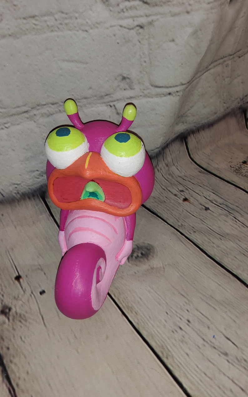 Op de afbeelding: Een handbeschilderd keramiek beeldje van een roze en oranje cartoon-slak met grote groene ogen en een open mond. De slak is opgerold in een spiraalvorm.