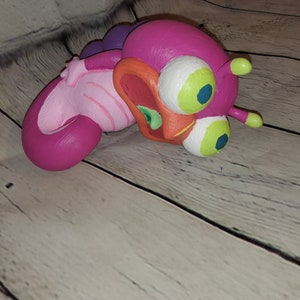Op de afbeelding: Een roze en paarse cartoonfiguur met grote groene ogen en een open mond. Het figuur is gemaakt van klei en heeft een getextureerd oppervlak.