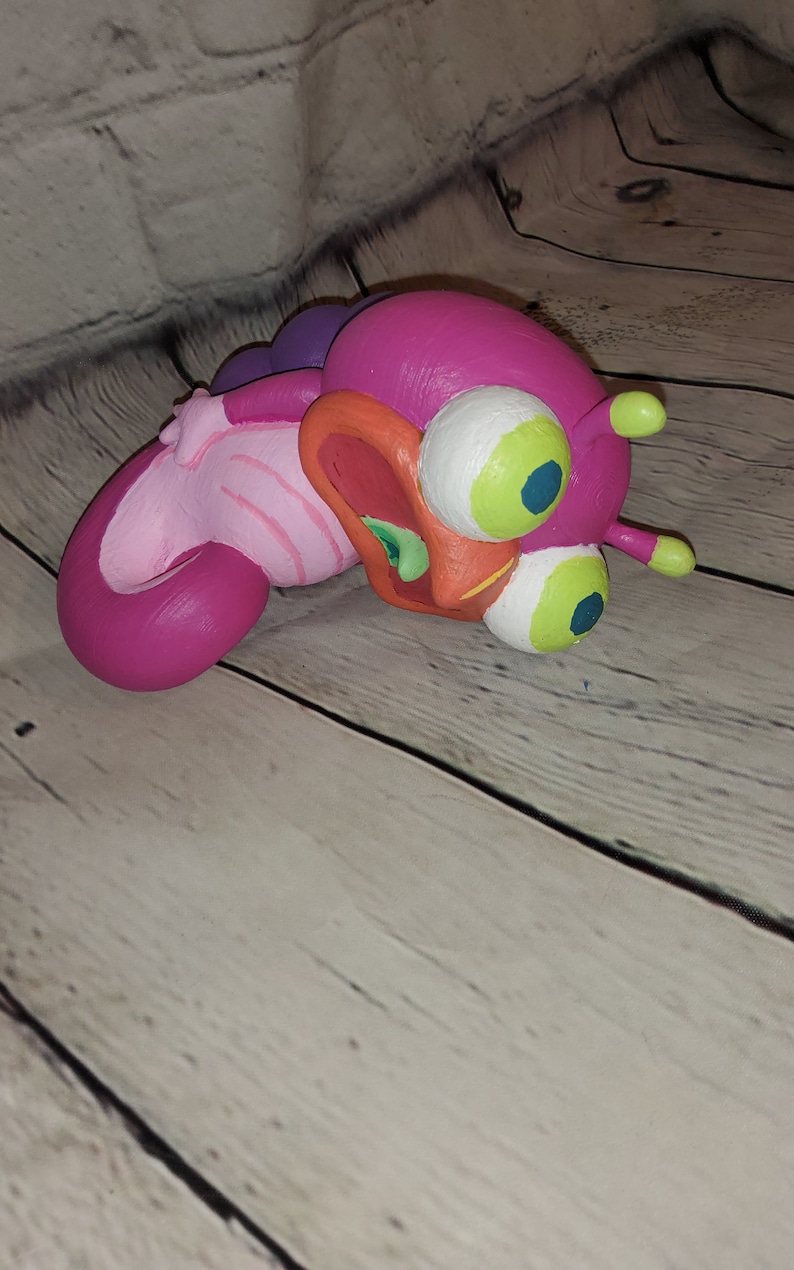 Op de afbeelding: Een roze en paarse cartoonfiguur met grote groene ogen en een open mond. Het figuur is gemaakt van klei en heeft een getextureerd oppervlak.